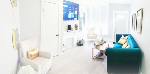Una sala de estar luminosa con un sofá blanco, un sillón verde azulado y una mesa de centro. Un televisor de pantalla plana está montado en la pared, y hay cuadros enmarcados.