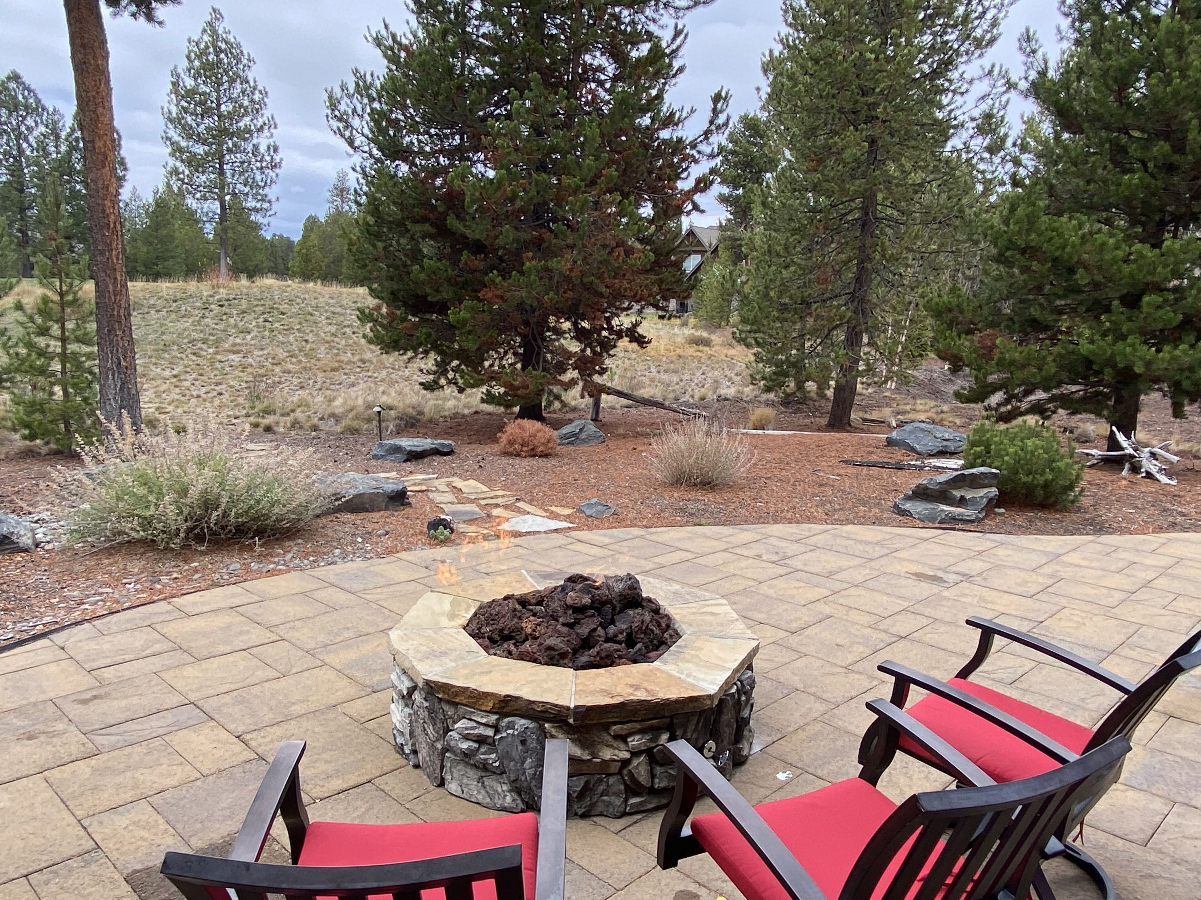 A patio with a stone fire pit, surrounded by chairs and a large tree.