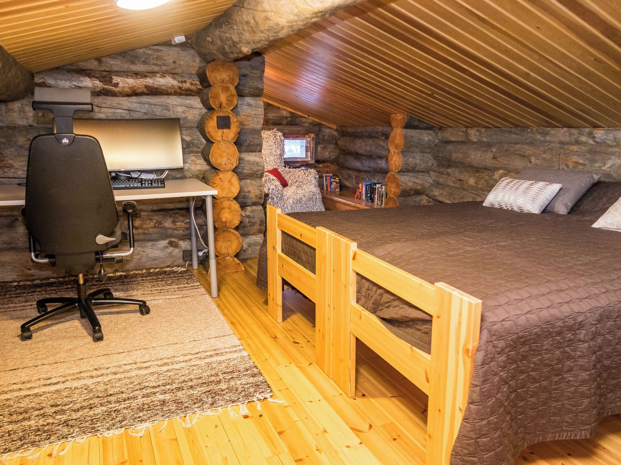 A cozy attic bedroom with a wooden desk, a computer, a chair, a bed with a quilt, and a small TV.