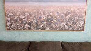 A room with a large framed landscape painting on the wall, a brown sofa with two cushions, and a textured light blue wall.