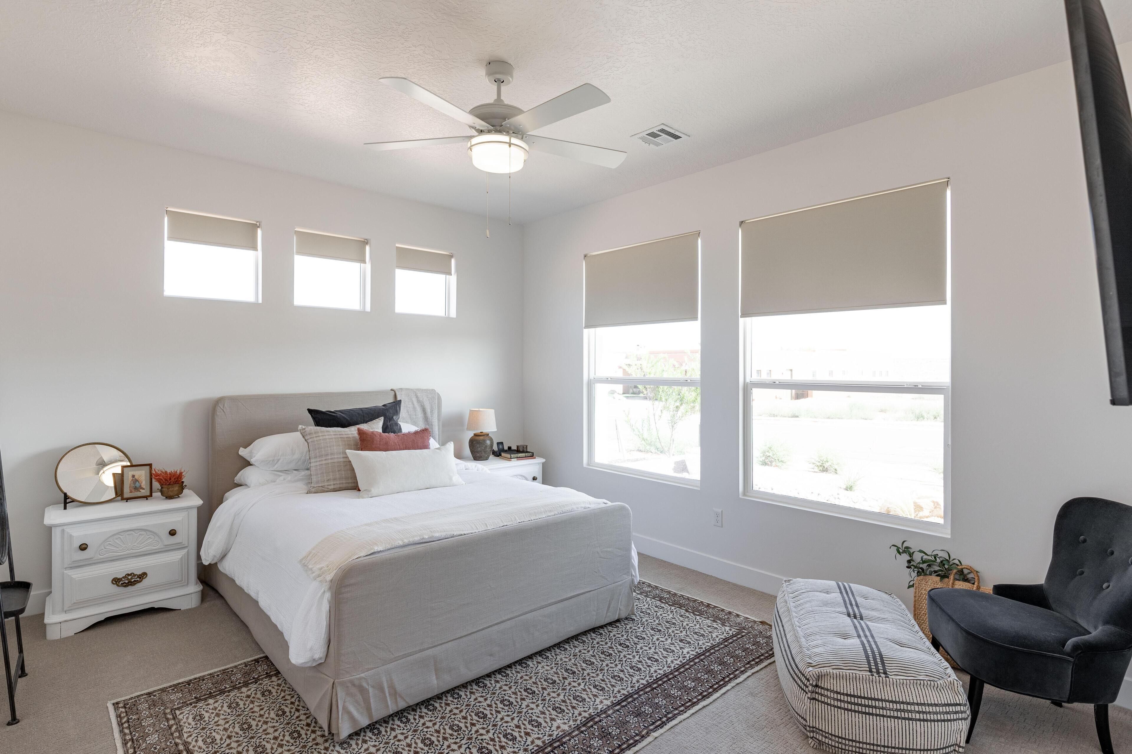 A bedroom with a bed, bedside tables, a chair, a ceiling fan, and large windows.