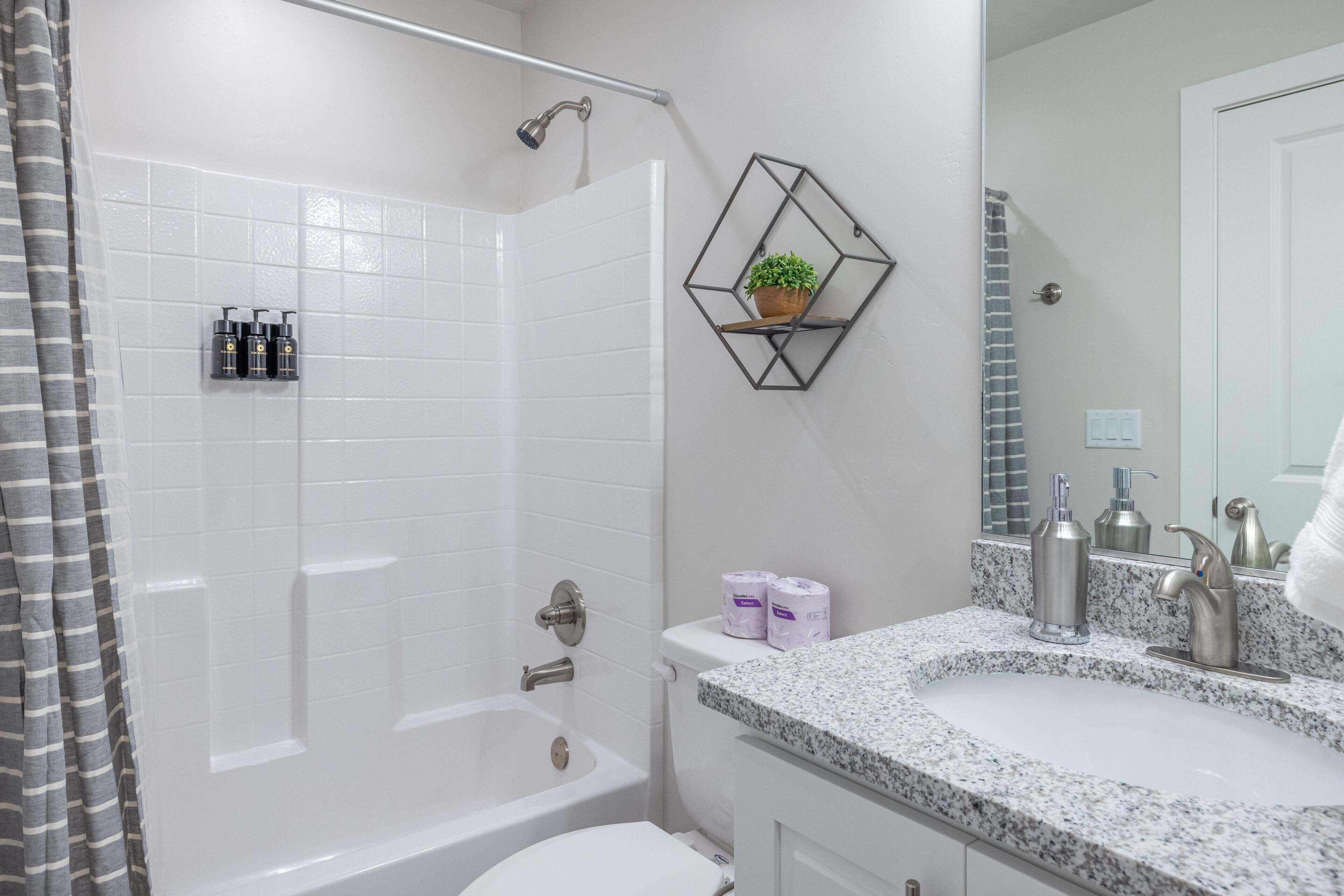 Une salle de bain avec une baignoire blanche, une douche, un lavabo avec un plan de travail en granit et un miroir. Un porte-serviettes avec une serviette et une plante en pot sont fixés au mur.