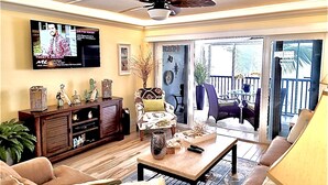 A living room with a television, sofa, coffee table, and a balcony with outdoor furniture. - Steps from Beach, Gorgeous Sunset Views, Recently Renovated, 2BD/2BTH, Sleeps 4 (Fort Myers Beach)