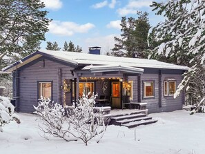 Lumipeitteinen mökki, jonka edessä on kuisti ulkokalusteineen ja jonka yllä on kirkas taivas.