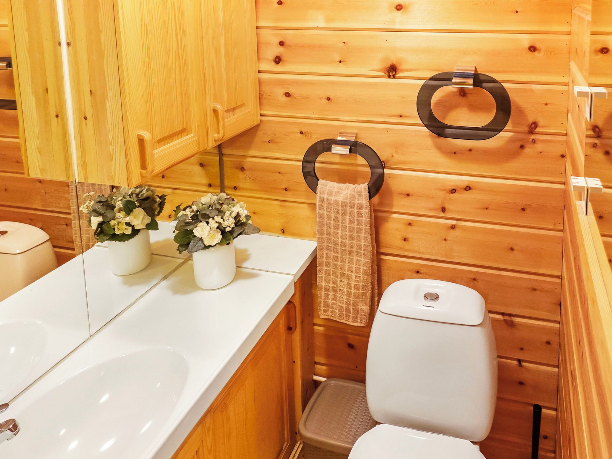 A wooden cabin-style bathroom with a white sink, two flower vases, a towel ring, and a toilet.
