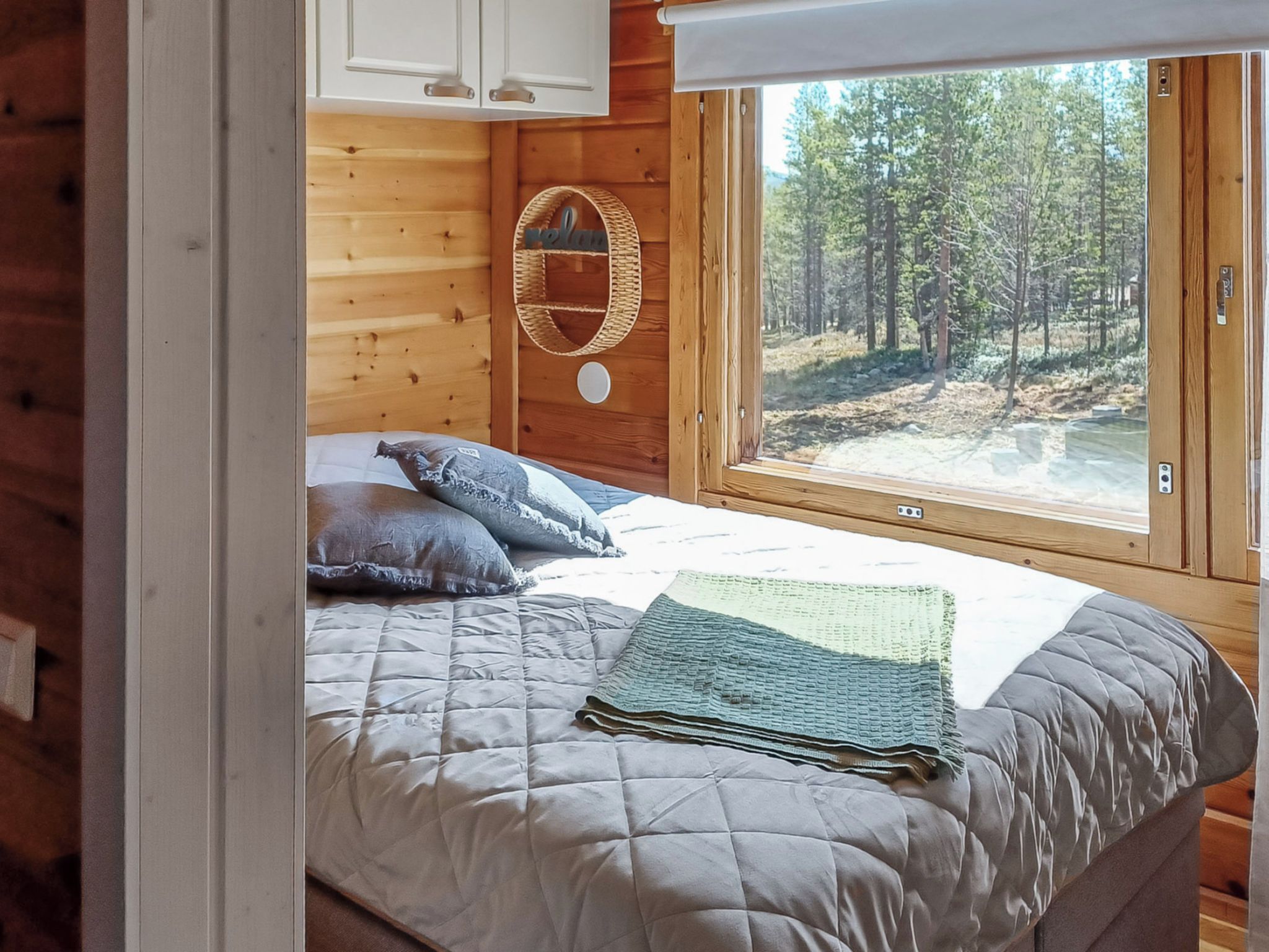 Une chambre à coucher dans une cabane en bois, avec un lit, des oreillers, un panier en osier et une fenêtre donnant sur une forêt.