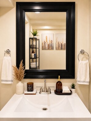 A bathroom with a large mirror, a white sink, a black shelf with toiletries, and two towels hanging on towel rings. - Chic Spacious 2BR Home Near Yonge St (Newmarket)