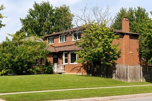 A two-story brick house with a front porch, a fenced yard, and trees. - Chic Spacious 2BR Home Near Yonge St (Newmarket)