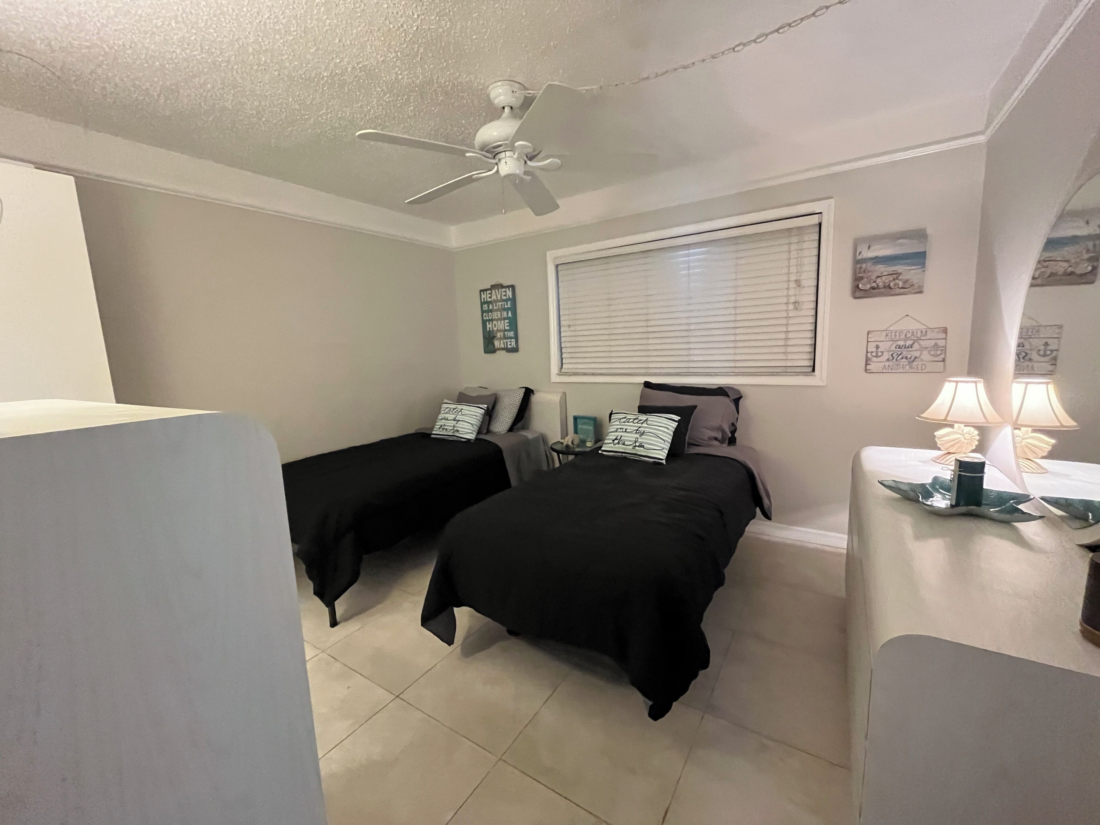 A bedroom with two beds, a ceiling fan, a window with blinds, and a bedside table with lamps.