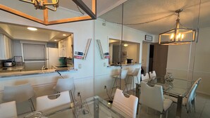 A modern kitchen with white cabinets, a built-in oven, and a stainless steel refrigerator. There is a dining area with a glass table and white chairs. - Amazing Beachfront Condo (Jensen Beach)