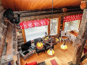 A rustic cabin interior with a wooden ceiling, a spinning wheel, a sofa, a dining table, and a fireplace.