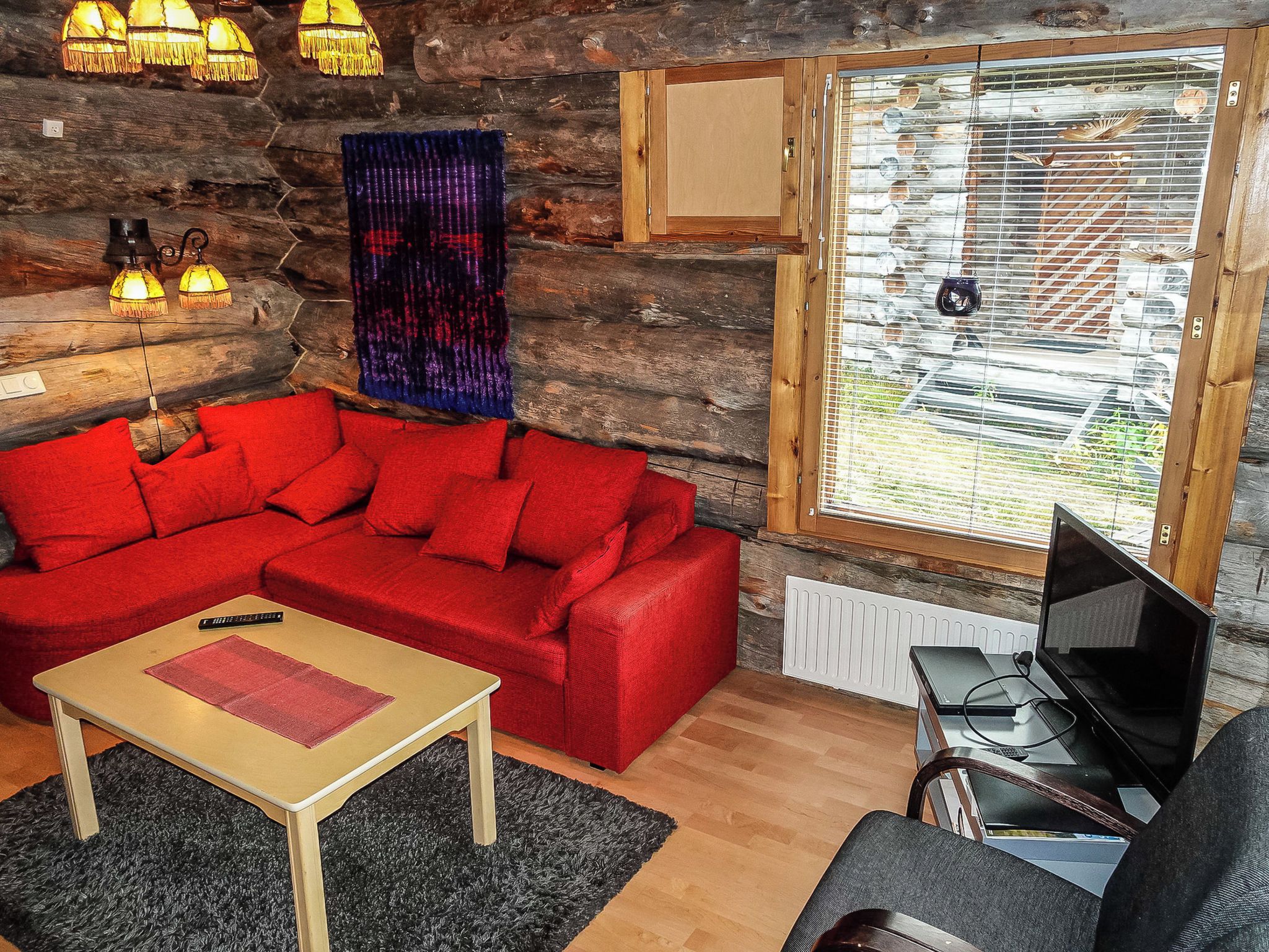 A cozy room with a red sofa, a small wooden table, a TV, and a window with blinds.