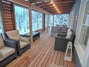 A covered outdoor deck with wooden flooring, wicker furniture, and large windows offering a view of snow-covered trees.