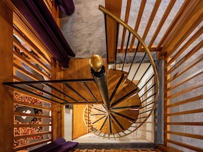 A spiral staircase with wooden railings and a golden handrail, leading to a carpeted landing area with a patterned rug and a purple cushion. - Bhakti Manor Creekside Home (Franklin)