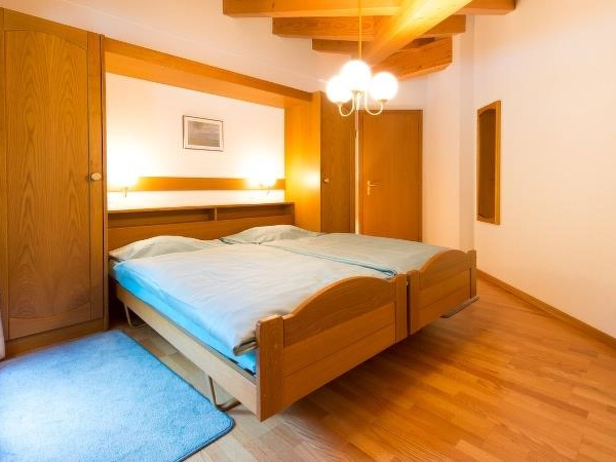 A bedroom with a wooden bed, a nightstand, a bedside table, a wall-mounted lamp, and a small picture on the wall.