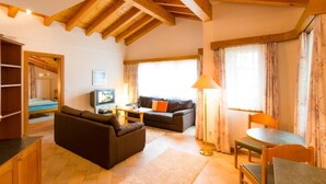 Ein gemütliches Wohnzimmer mit einer Couch, einem kleinen Tisch und einem Fernseher. Die Decke ist aus Holz und es gibt ein Fenster mit Vorhängen.