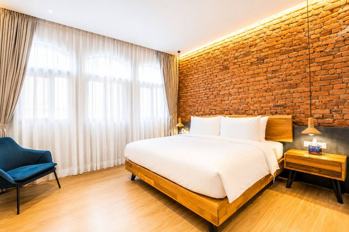 A hotel room with a large bed, a blue chair, a wooden nightstand, and a brick wall.