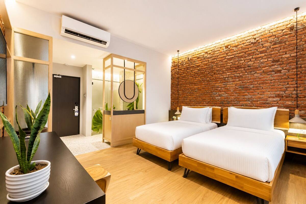 A modern hotel room with two beds, a brick accent wall, a wooden desk, and a potted plant.