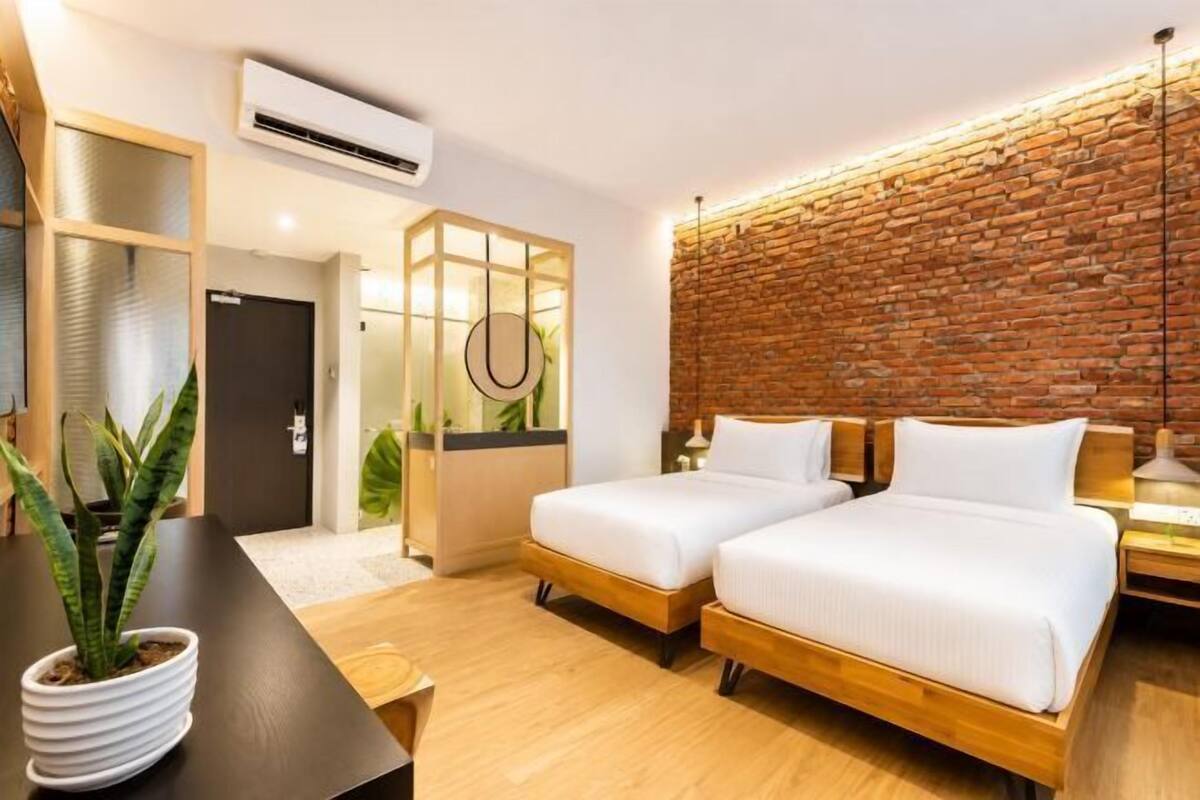 A hotel room with two beds, a wooden floor, a brick wall, and a potted plant.