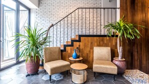 A modern interior with two beige armchairs, a round wooden table, and potted plants. A staircase with a black railing leads to a wooden wall. - Tien Terrace (George Town)