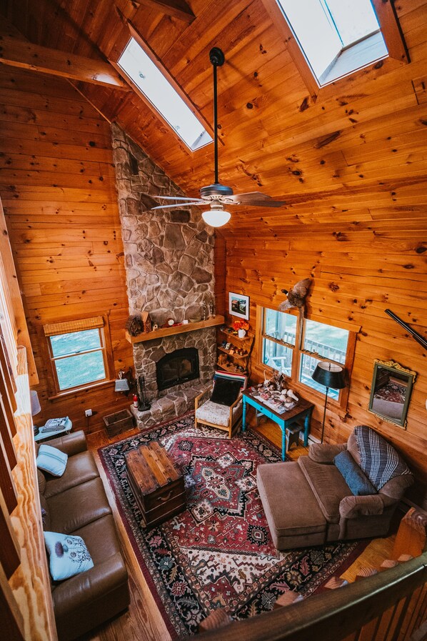 Ein gemütliches Blockhaus-Interieur mit Kamin aus Stein, Holzwänden, Deckenventilator und großem Fenster für natürliches Licht.