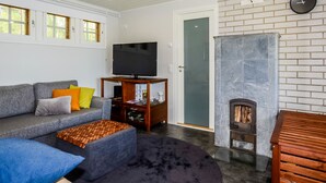 Een woonkamer met een grijze bank, een zwart tapijt, een houten dressoir, een open haard en een tv op een standaard.