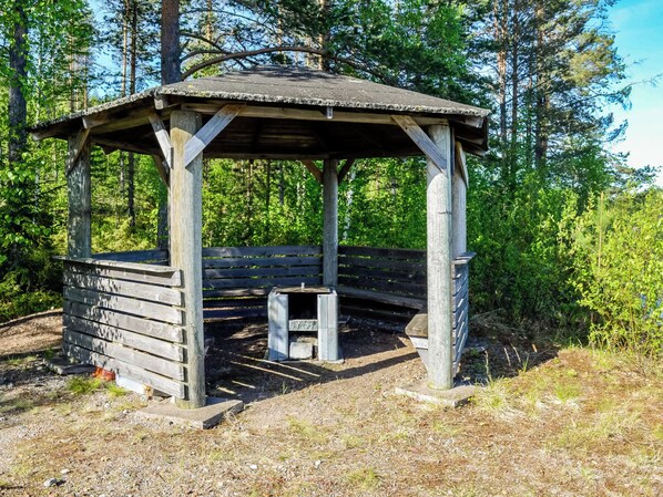 En trälusthus med en bänk och en eldstad i ett skogsområde.