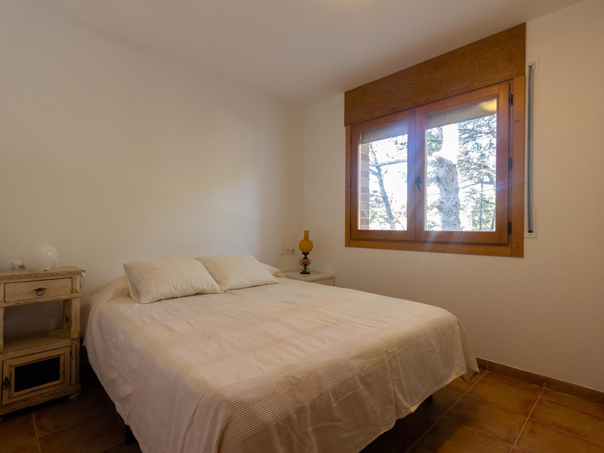 Un dormitorio con cama, mesita de noche, lámpara y ventana con vista a árboles.