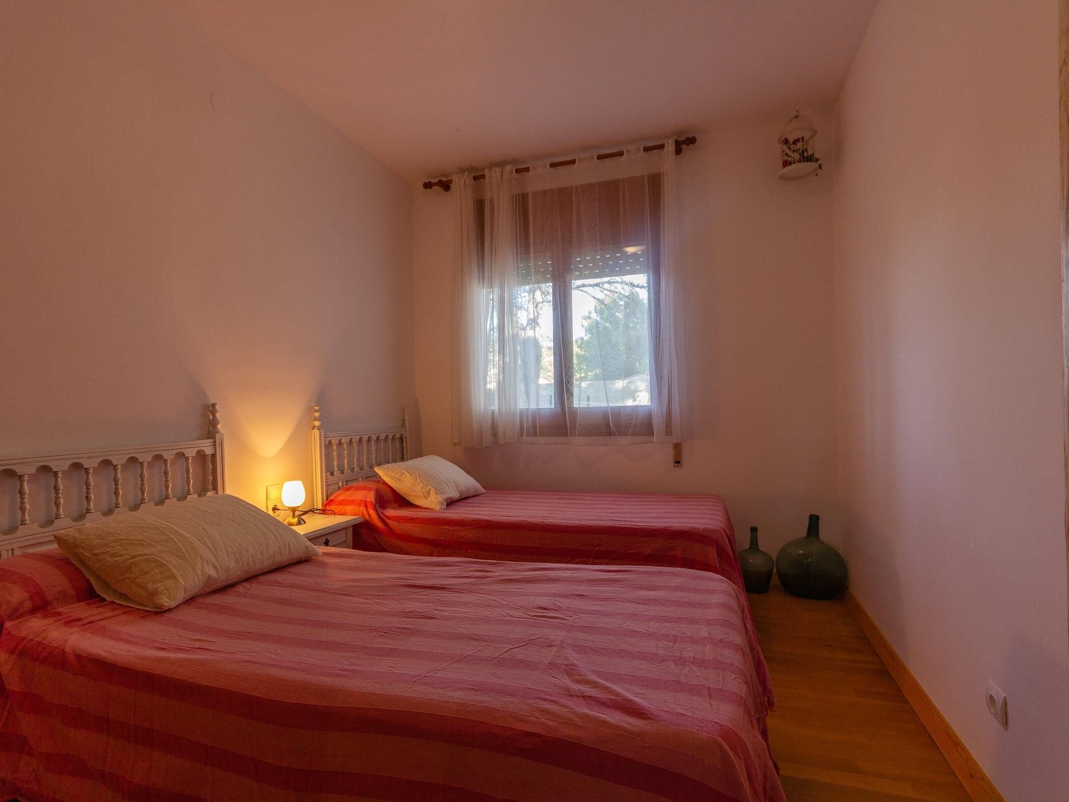 Una habitación con dos camas, una mesita de noche con lámpara, una ventana con cortinas traslúcidas y un pequeño objeto decorativo en la pared.
