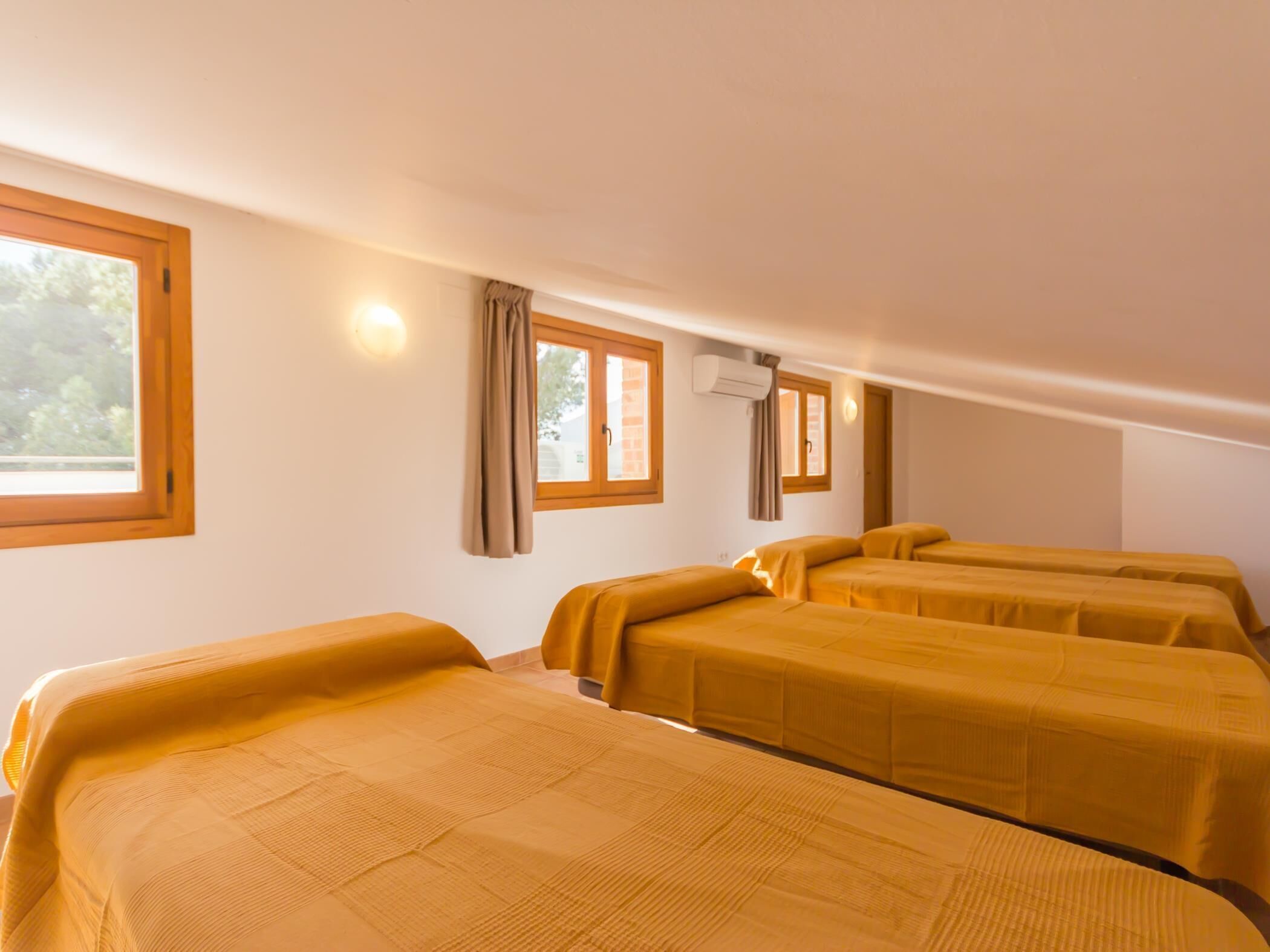 A room with three beds, each with a headboard and a window. There is an air conditioning unit on the wall.