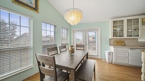 A dining room with a wooden table, chairs, a chandelier, and a view through large windows with blinds. - Ocean View, Short Walk to Beach, Free Golf Cart Included (Port Aransas)