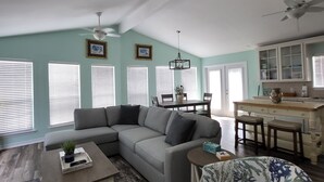 A spacious living room with a sectional sofa, a coffee table, and a patterned armchair. There is a dining area with a table and chairs, and a kitchen with cabinets and a countertop. - Ocean View, Short Walk to Beach, Free Golf Cart Included (Port Aransas)