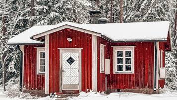 Eine rote Holzhütte mit weißer Tür und Fenstern, umgeben von schneebedeckten Bäumen.
