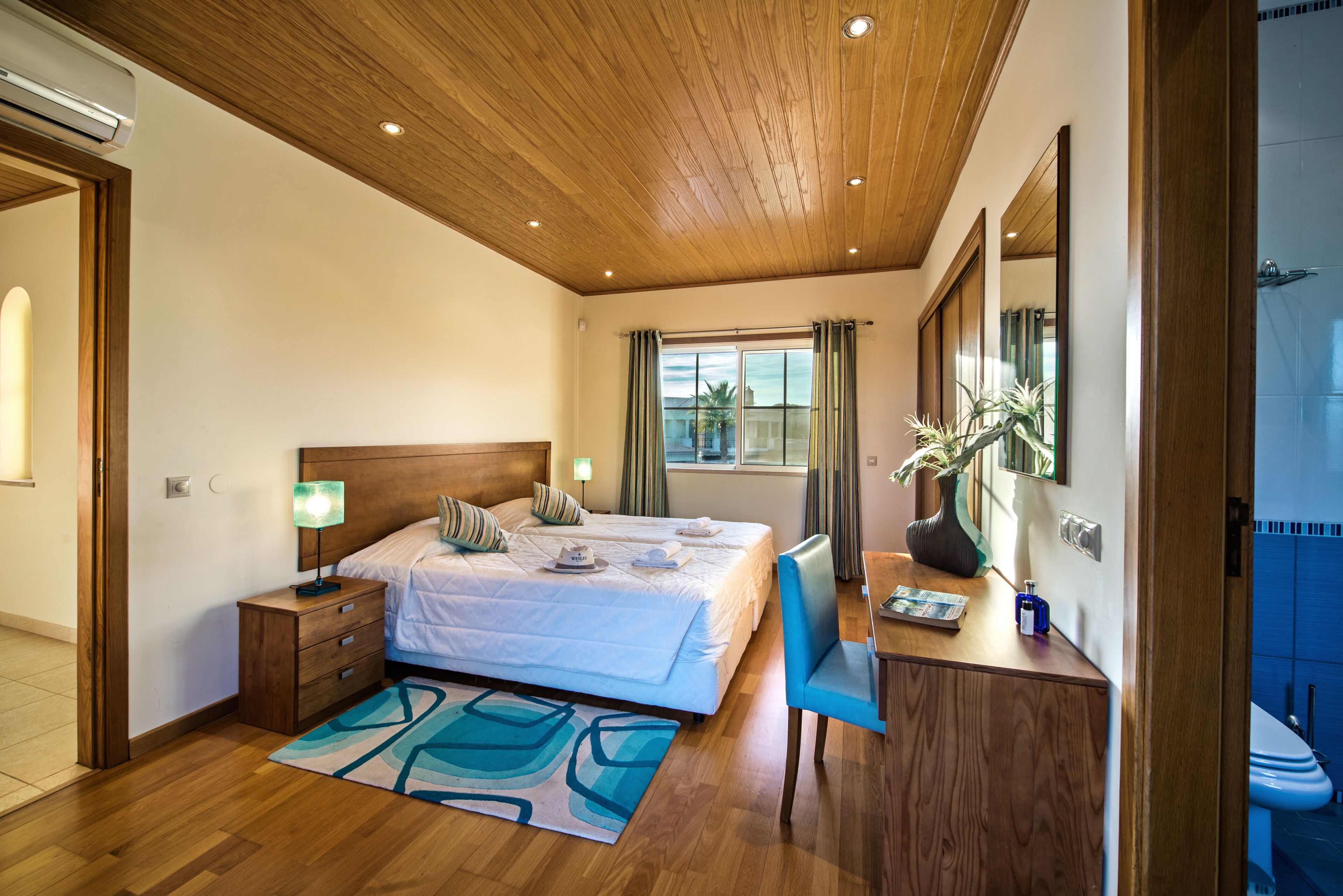 Une chambre moderne avec un plafond en bois, un grand lit, une table de chevet, un bureau avec une chaise et une salle de bain visible à travers une porte ouverte.