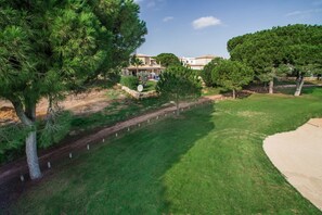 Stroll through the beautifully landscaped grounds, featuring a mix of pine and deciduous trees, along a paved pathway to a single-story house with a welcoming porch.