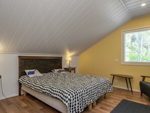 A bedroom with a sloped ceiling, a bed with a patterned bedspread, a bedside lamp, a small table, and a window with blinds. - Isokoskelo by Interhome (Pertunmaa)