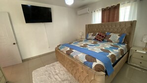 Ein Schlafzimmer mit Bett, einem an der Wand montierten Fernseher, einem Nachttisch mit Lampe und einem Vorhangfenster.