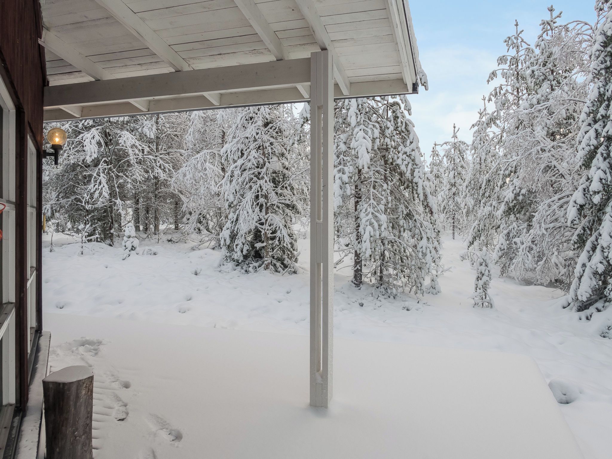 Schneebedeckte Landschaft mit einer Holzhütte, vermutlich einem Winterbalkon, und mit Schnee beladenen Bäumen.