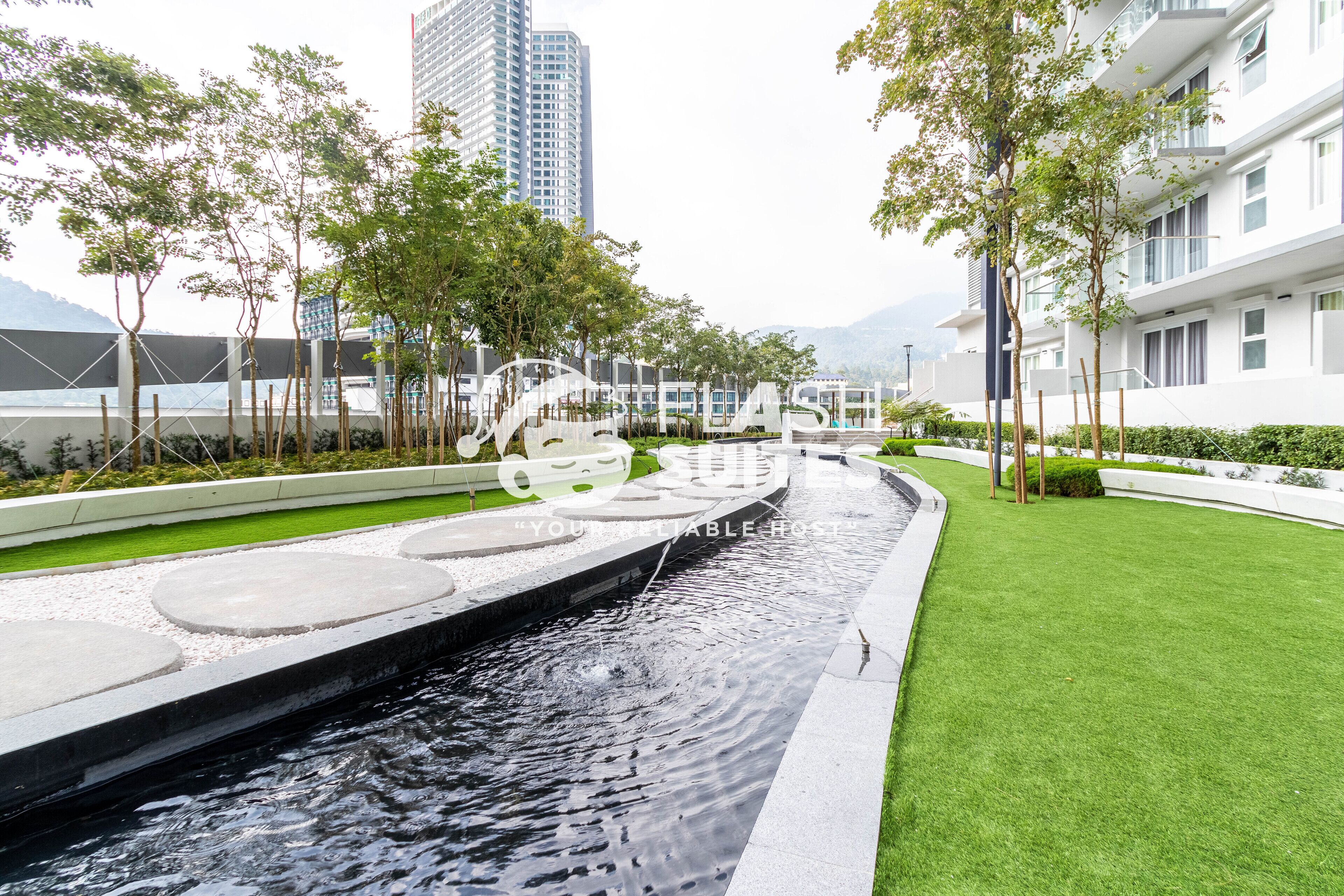 A modern hotel with a water feature, green lawns, and a view of a tall building in the background.