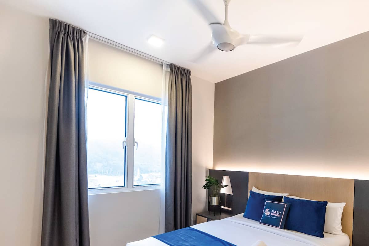 Une chambre d’hôtel avec un lit, une table de chevet, une lampe, un ventilateur de plafond et une fenêtre avec des rideaux.