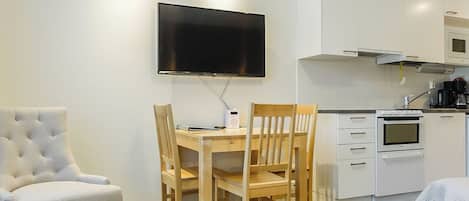 A modern living room with a flat-screen TV mounted on the wall, a dining table with chairs, a sofa, and a kitchenette with a microwave and oven.