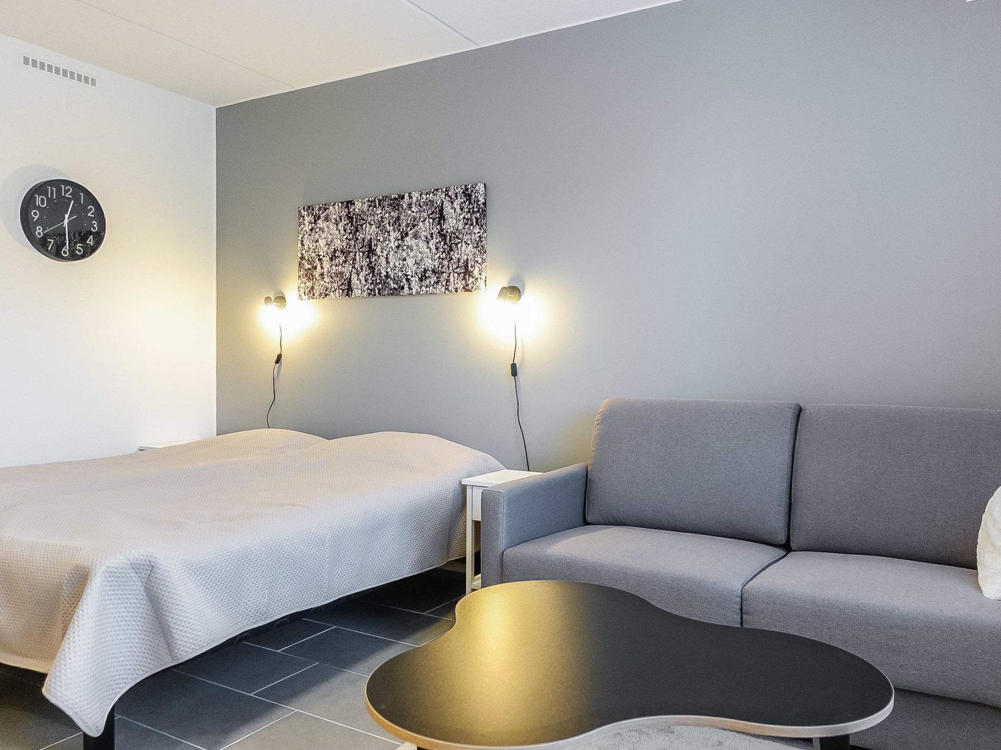 Et moderne hotelværelse med seng, grå sofa, lille bord og vægur.