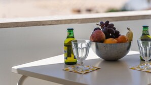 Uma mesa com uma tigela de frutas, duas garrafas e dois copos numa varanda com vista para a praia.