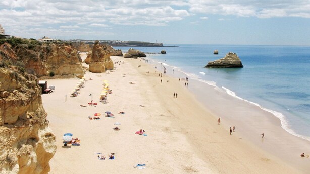 Uma praia com falésias rochosas, guarda-sóis e pessoas.