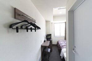 Une chambre d’hôtel compacte comprenant un lit, un bureau avec un ordinateur, une télévision et un porte-manteau fixé au mur.