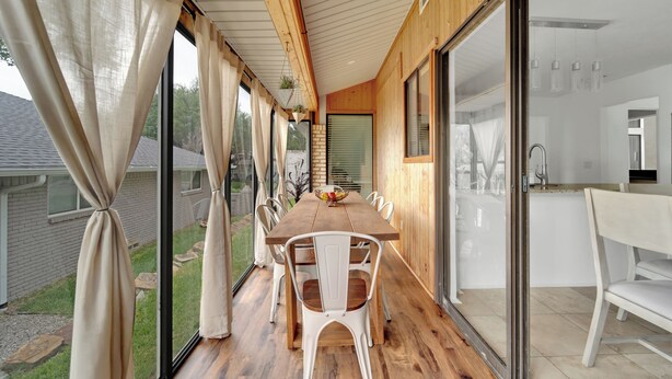 A wooden deck with a dining table and chairs, a kitchen area with a sink and cabinets, and large sliding glass doors leading to a garden.