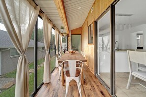 Une terrasse en bois avec une table et des chaises, un coin cuisine équipé d’un évier et de placards, et de grandes portes-fenêtres coulissantes donnant sur un jardin.