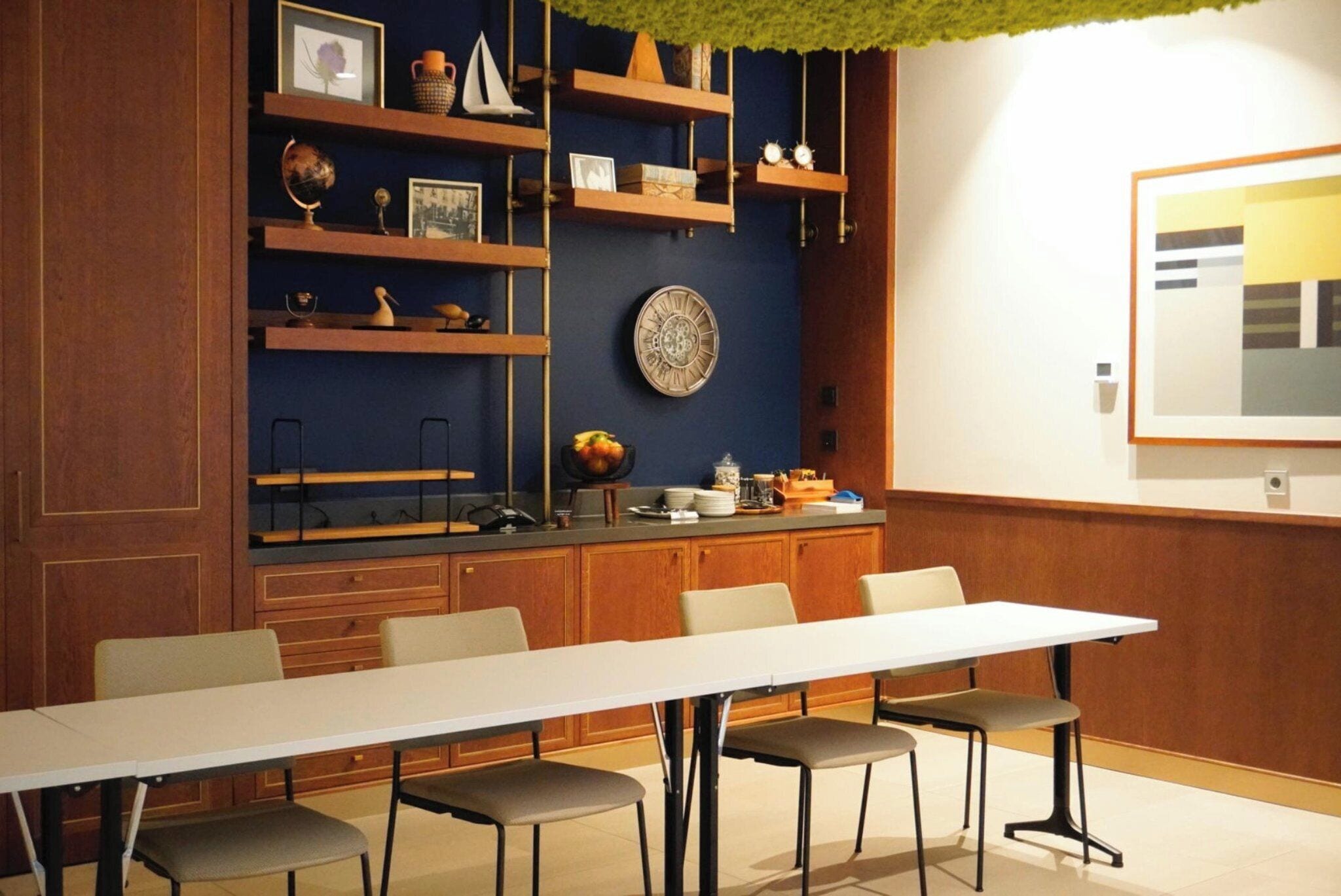 Une salle de conférence avec une longue table et quatre chaises, des étagères en bois où sont rangés divers objets, et une horloge au mur.