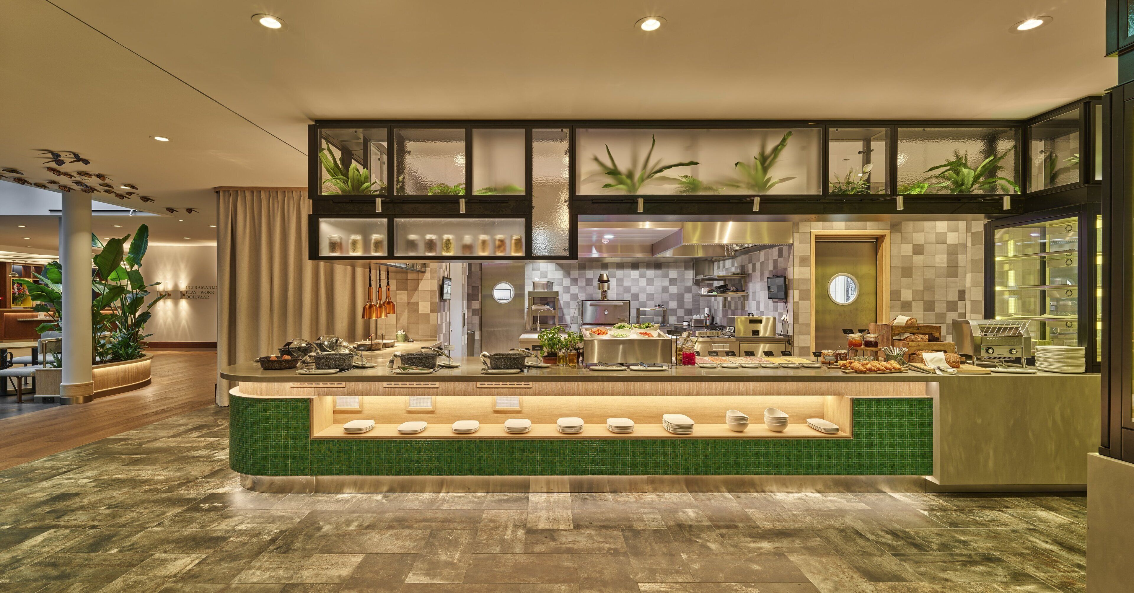 A modern hotel lobby with a reception desk, a dining area, and a kitchen in the background.