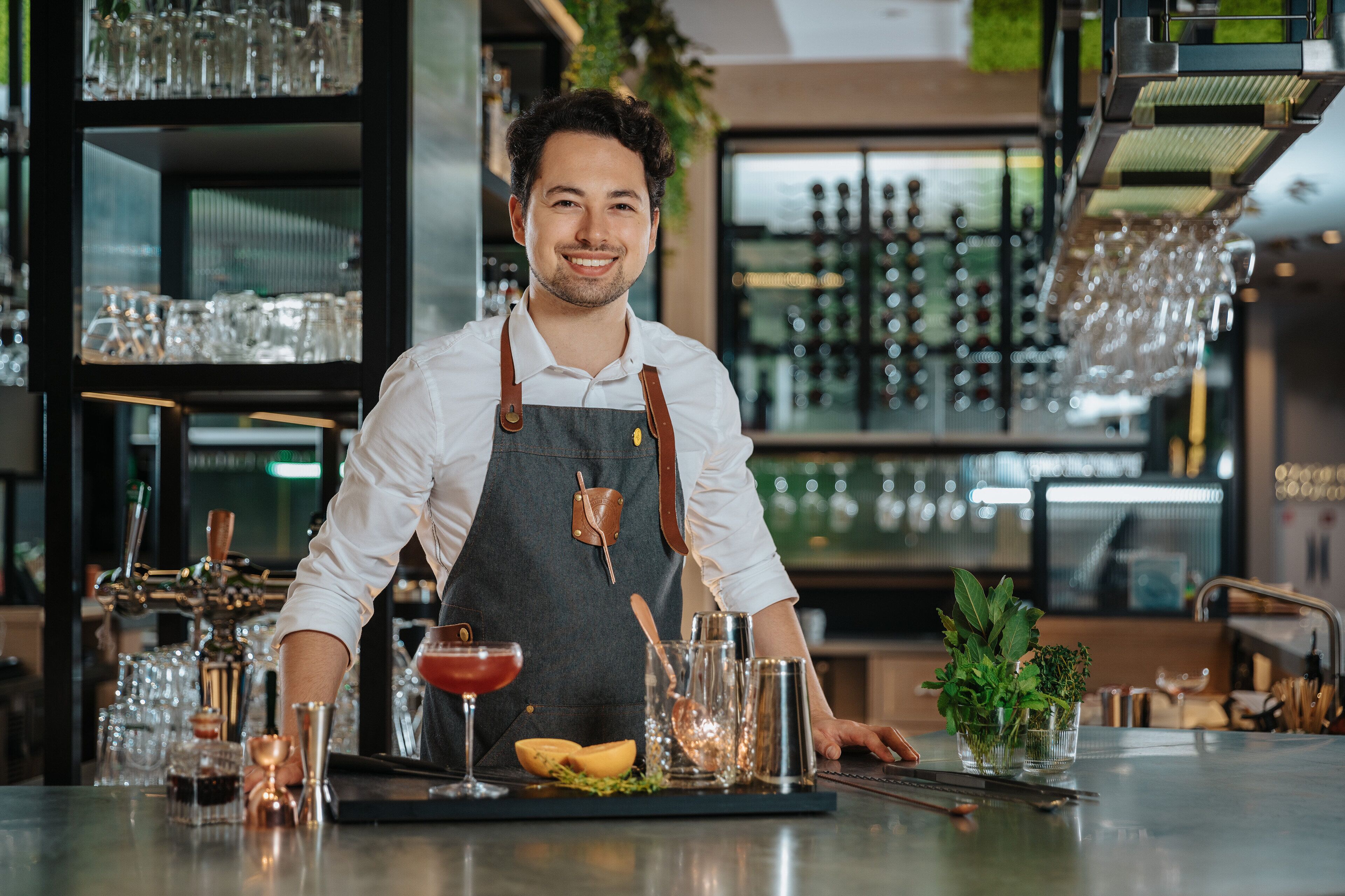 A bartender at a bar with a well-stocked shelf of glasses and bottles, a cocktail on the counter, and a variety of bar tools.